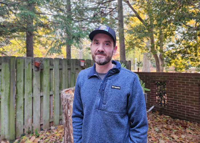 man wearing a blue quarter zip sweater and a blue hat posing in front of a green fence